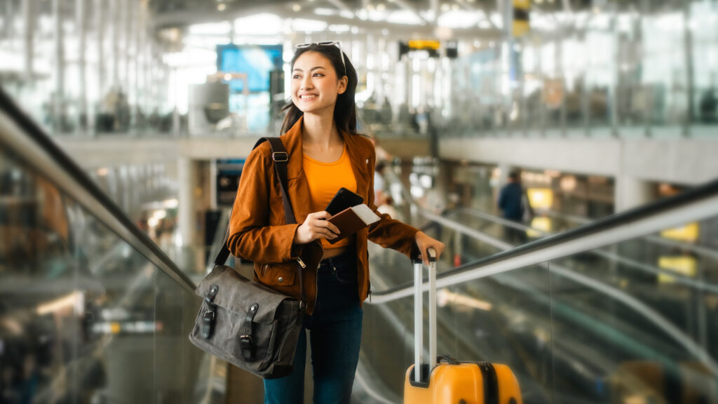 happy young asian woman traveler holding boarding pass ticket, passport and smartphone at the airport terminal with her luggage and smiling at camera, cheerful tourist female having holiday trip
